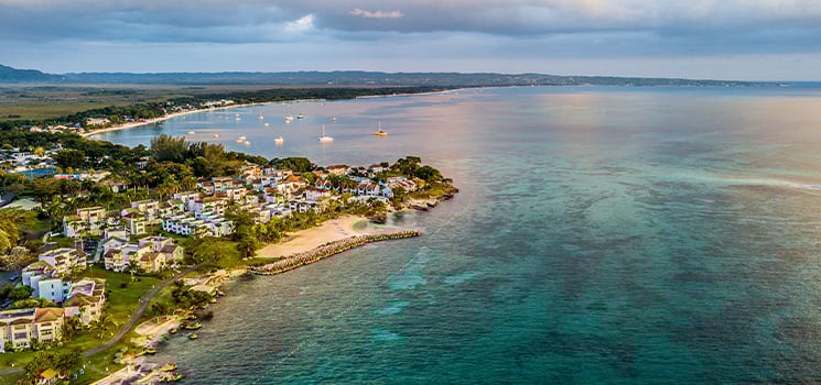 Aerial view of a serene coastal village in Jamaica, featuring calm turquoise waters, luxurious villas, and sailboats anchored in the bay under a cloudy evening sky.