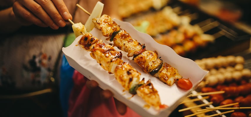 Hand holding grilled skewers with glazed sauce on white paper at Thai street food market.