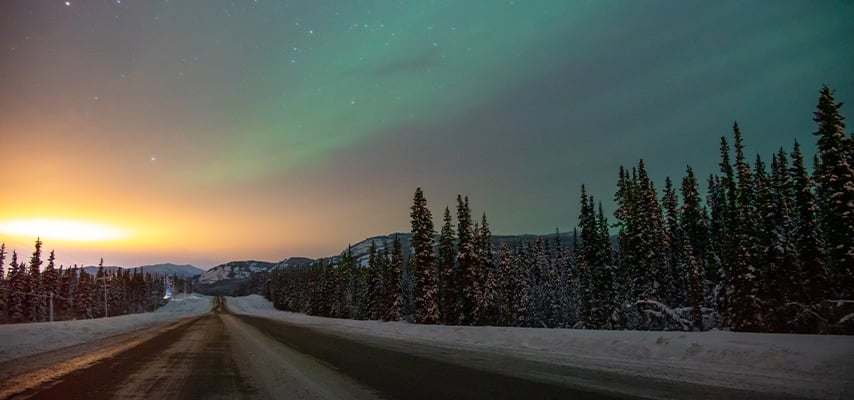 Northern lights glowing green over snowy Alaskan road with evergreen trees at dusk