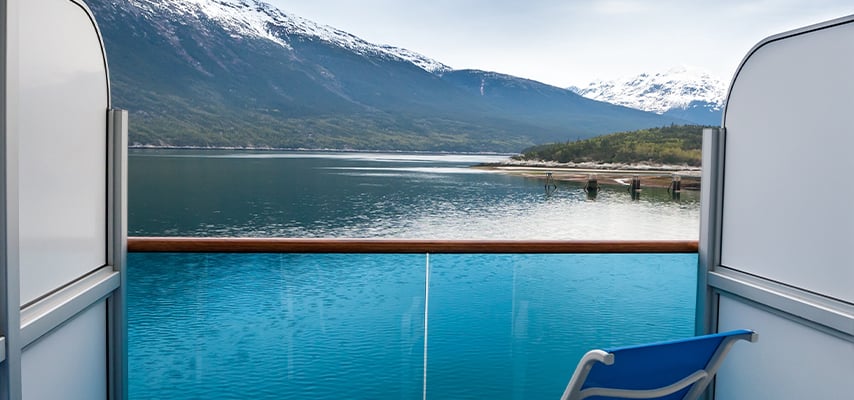 View from Alaskan cruise ship balcony showing snow-capped mountains reflected in calm blue water with wooden dock.