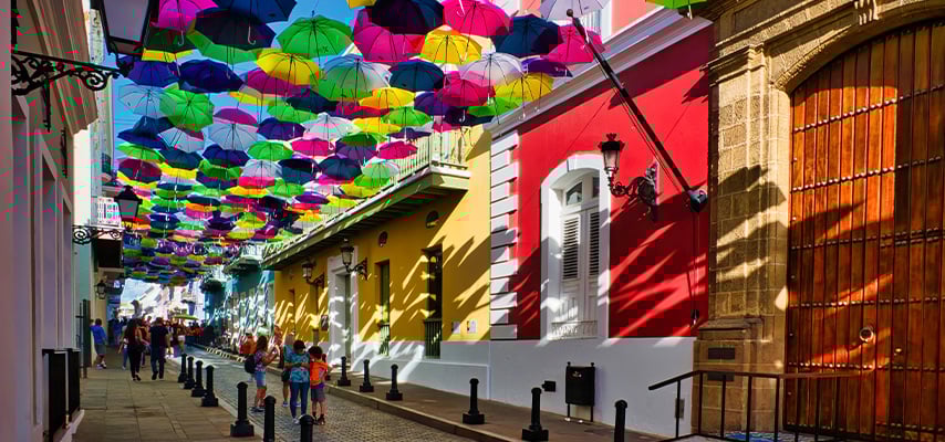Colorful umbrella art installation in Old San Juan, Puerto Rico, with historic colonial architecture featuring yellow and red buildings along Fortaleza Street during peak tourist season.