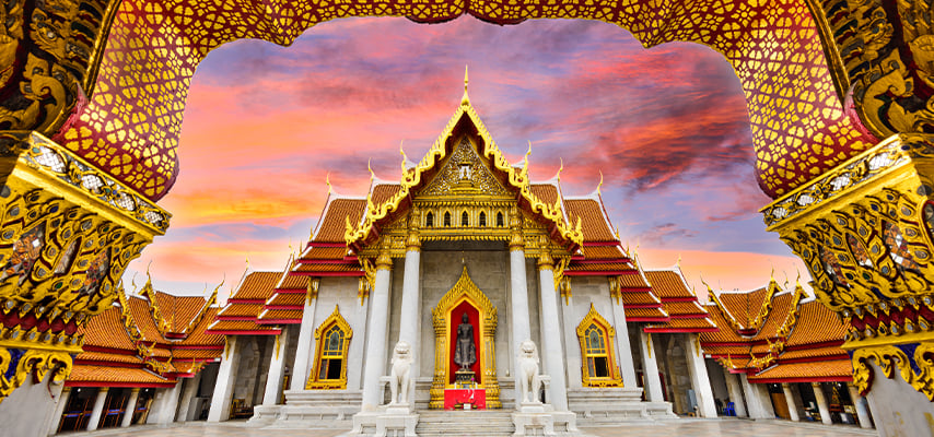 Ornate Thai temple with golden spires and white walls framed by decorative golden archway at sunset.