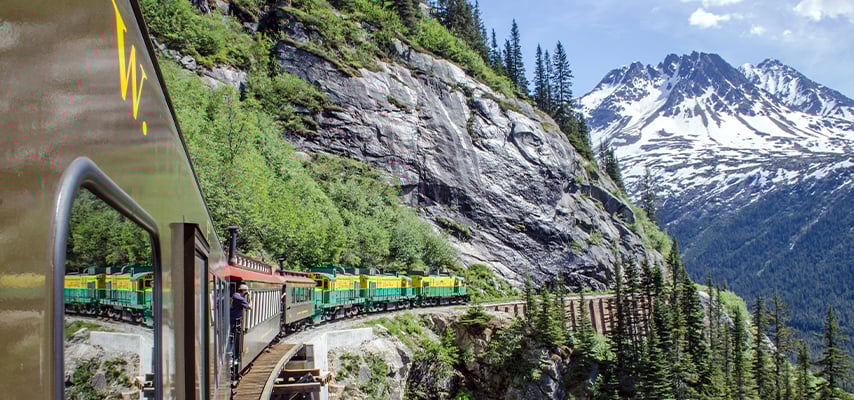 White Pass &amp; Yukon Railway train winding through Alaskan mountains with snow-capped peaks and rocky cliffs.