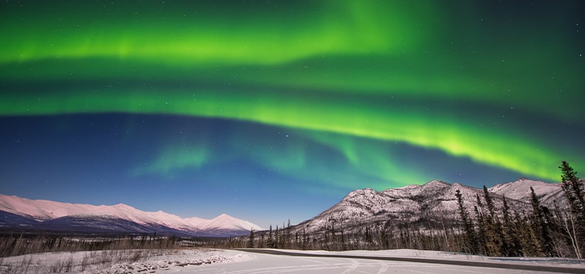Brilliant green northern lights streaming across sky above snowy mountain highway in Alaska