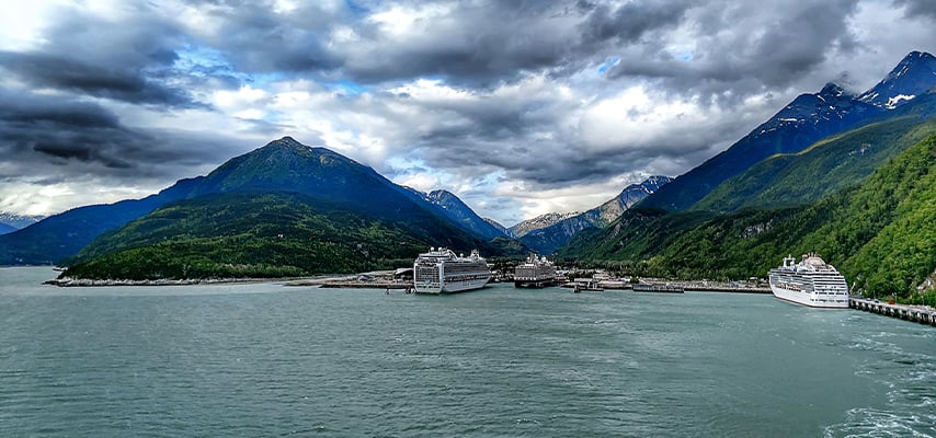 Cruise ships docked at Alaskan port surrounded by dramatic mountains under stormy cloudy skies.