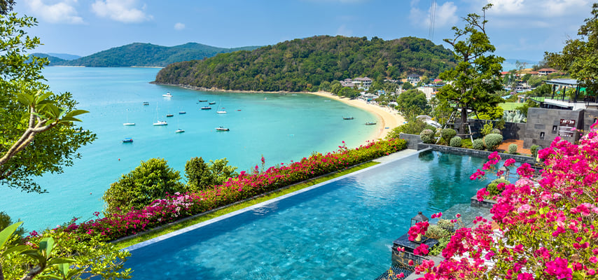 Infinity pool overlooking turquoise bay with boats and forested hills, bordered by vibrant bougainvillea flowers.