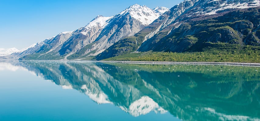 Snow-capped Alaskan mountains perfectly reflected in still turquoise water on a clear sunny day.