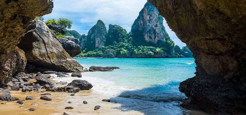 Turquoise water and limestone cliffs viewed through rocky cave opening at tropical Thai beach.