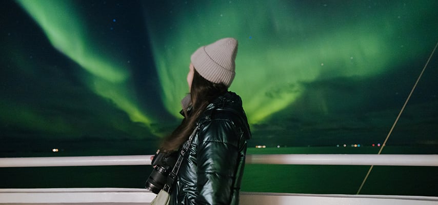 Side view of happy female traveller with camera overlooking dramatic bright Aurora in the sky while crossing the ocean by the ship in Northern Norway