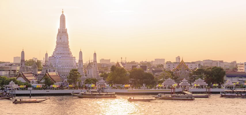 Wat Arun temple spire rises above Bangkok skyline at sunset with Chao Phraya River and boats.