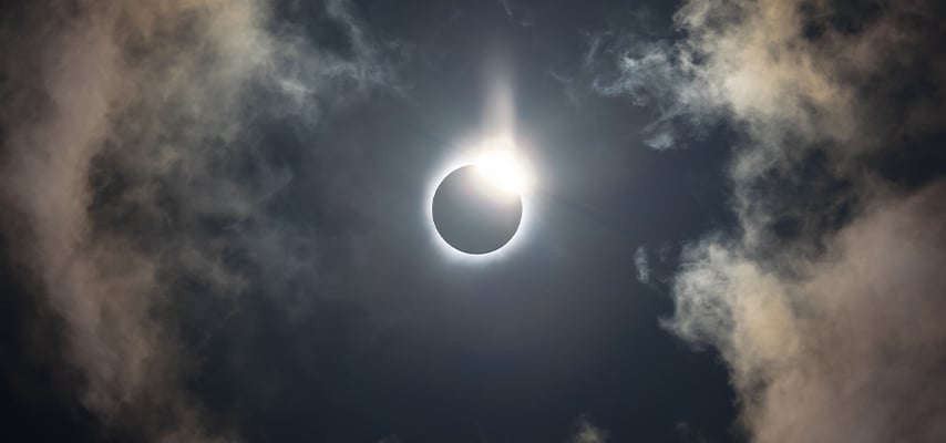 Total solar eclipse with bright corona and diamond ring effect visible through dramatic cloudy sky.