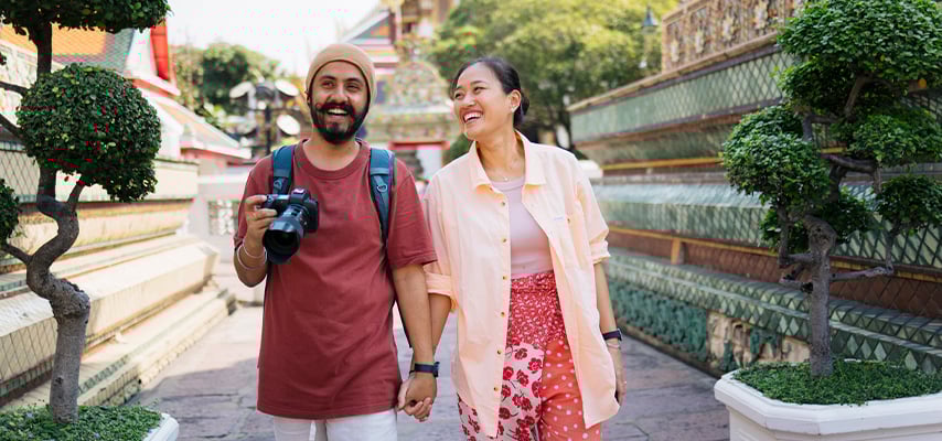 Happy couple holding hands and camera while exploring ornate Thai temple grounds together.