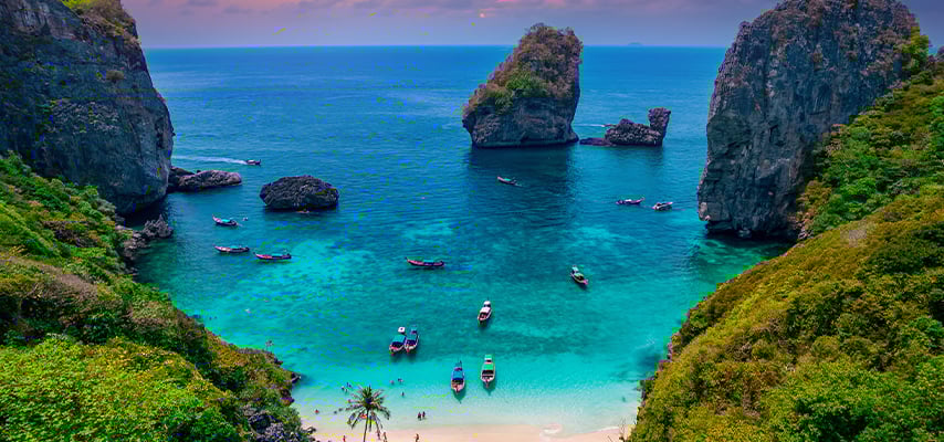Turquoise cove surrounded by dramatic limestone cliffs with longtail boats anchored near white sand beach.