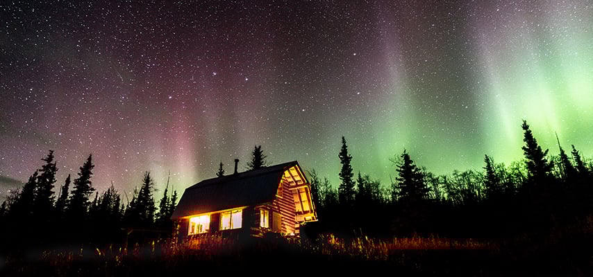 Illuminated log cabin under green and pink aurora borealis with evergreen trees in Alaska