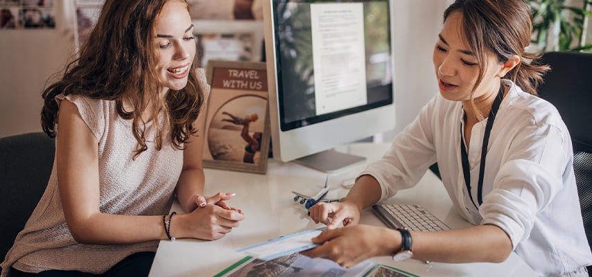 Travel agent consulting with a female client, reviewing documents and planning a trip together at a desk with computer.