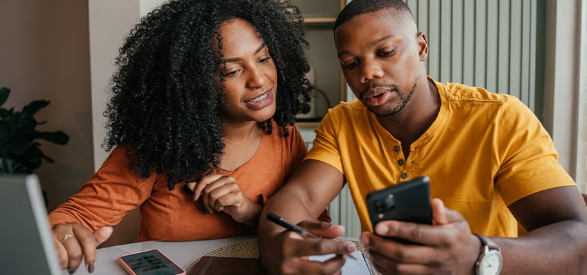 Couple sitting together at a table planning a trip, looking at smartphones and discussing travel options.