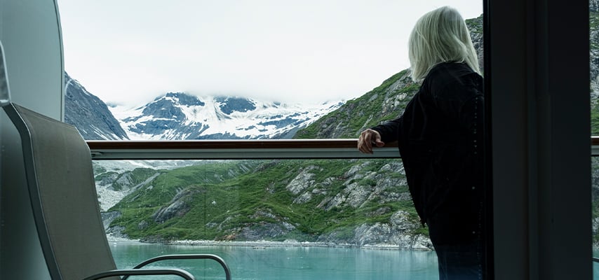 Woman looking out cruise ship window at snow-covered mountains and green coastline.