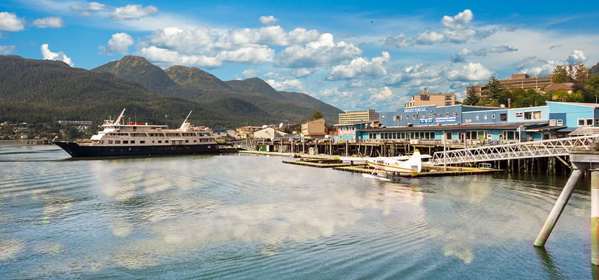 Small ferry and waterfront buildings reflected in calm harbor with mountains in background.
