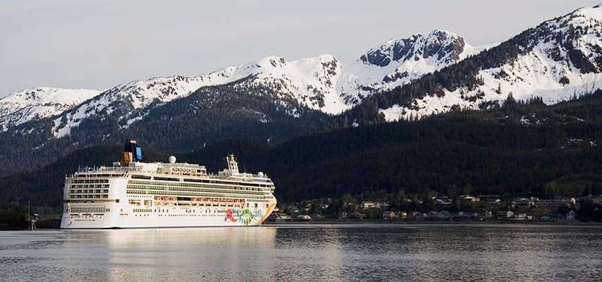 White cruise ship sailing past snow-covered mountains and forested Alaskan coastline.