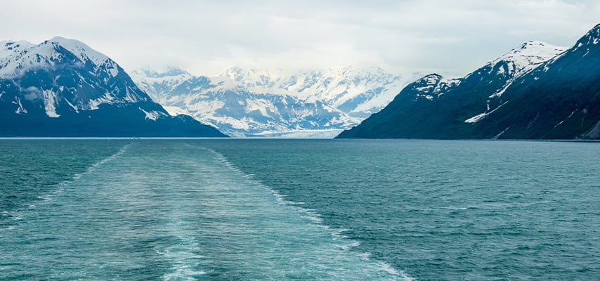 Ship's wake trailing through blue-green water between steep snow-capped mountain fjords.