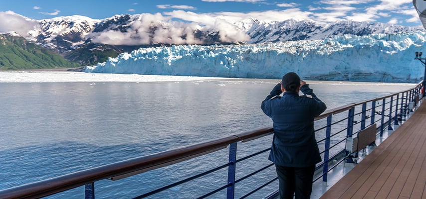 Person photographing massive blue glacier and mountains from cruise ship deck railing.