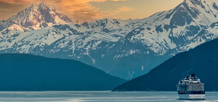 Cruise ship sailing through Alaskan waters at sunset with dramatic snow-covered peaks.