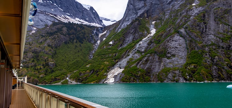 Cruise ship deck view of emerald waters and steep mountain cliffs along an Alaskan fjord.