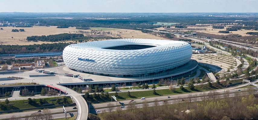 Aerial view of the Allianz Arena in Munich, its distinctive white diamond-paneled exterior surrounded by highways and open fields.