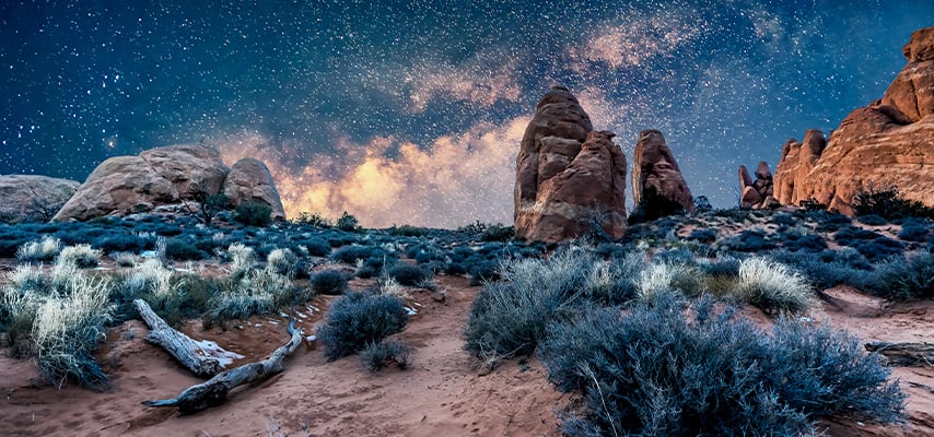 A starry night sky with the Milky Way glowing above red sandstone spires and desert shrubs at Arches National Park, Utah.