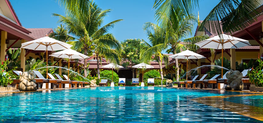 Tropical resort pool surrounded by palm trees, lounge chairs, and white umbrellas under blue sky.