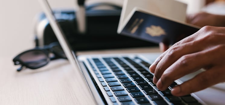 A person typing on a laptop keyboard with their left hand while holding their passport in their right hand.