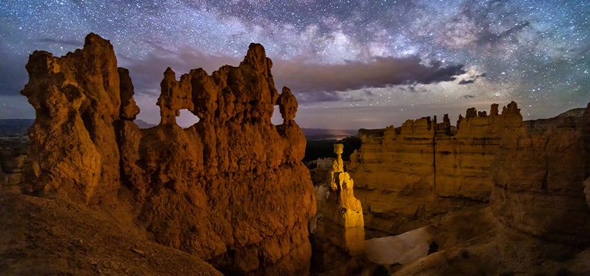 Illuminated orange hoodoo rock formations at Bryce Canyon National Park beneath a dramatic night sky scattered with stars and the Milky Way.