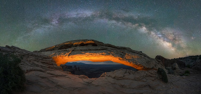 The Milky Way arches over Mesa Arch at Canyonlands National Park, with warm light glowing through the sandstone opening onto a canyon vista.
