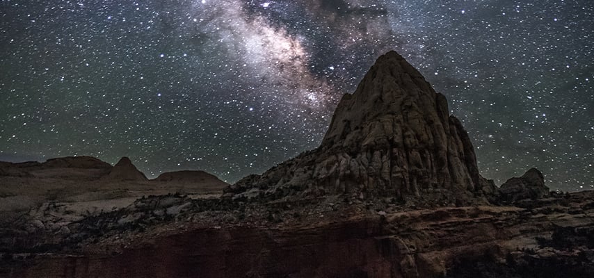 A towering sandstone butte rises against a brilliant star-filled night sky with the Milky Way visible at Capitol Reef National Park, Utah.