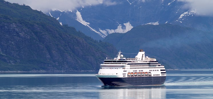 Large cruise ship sailing through calm Alaskan waters with snow-capped mountains in the background.