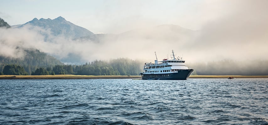 Small cruise ship navigating misty waters with forested Alaskan mountains shrouded in clouds.