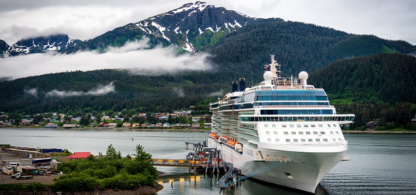 White cruise ship docked at Alaskan port with snow-capped mountains and small town visible.