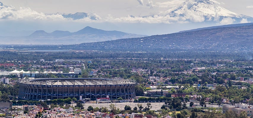 Aerial view of Estadio Azteca in Mexico City, nestled among dense urban sprawl with a snow-capped volcano visible in the distance.