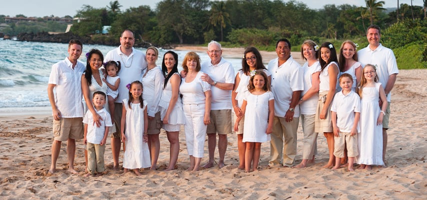 Large family in coordinating white and neutral outfits gathers on tropical beach for group portrait.