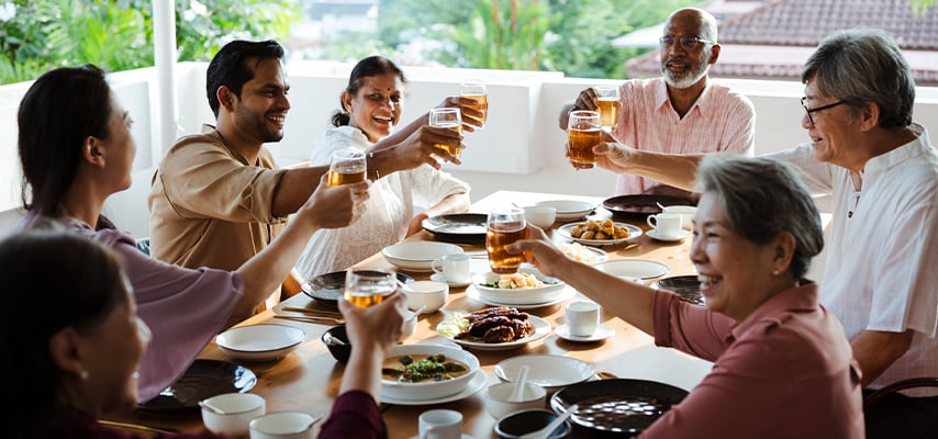 Family members toast with drinks around dining table filled with dishes at bright indoor gathering.