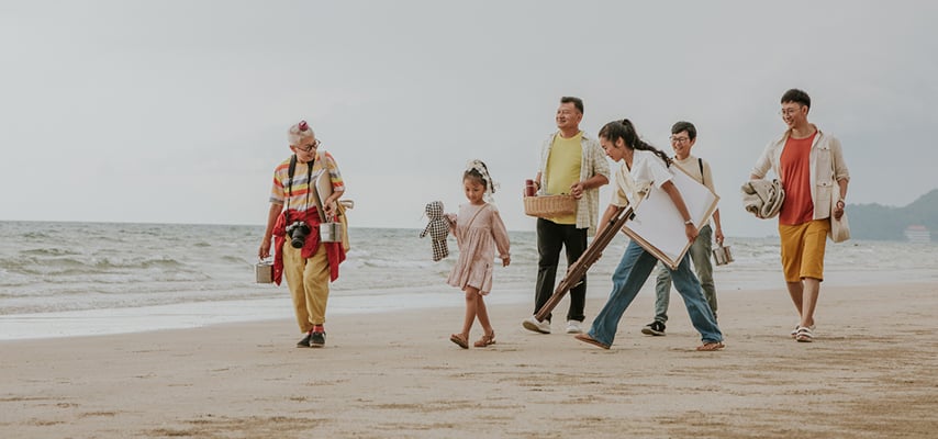 Multigenerational family walks along beach at sunset carrying blankets and beach gear together.