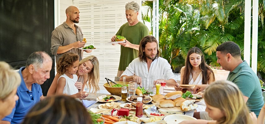 Extended family shares meal at outdoor table surrounded by tropical plants on sunny patio.