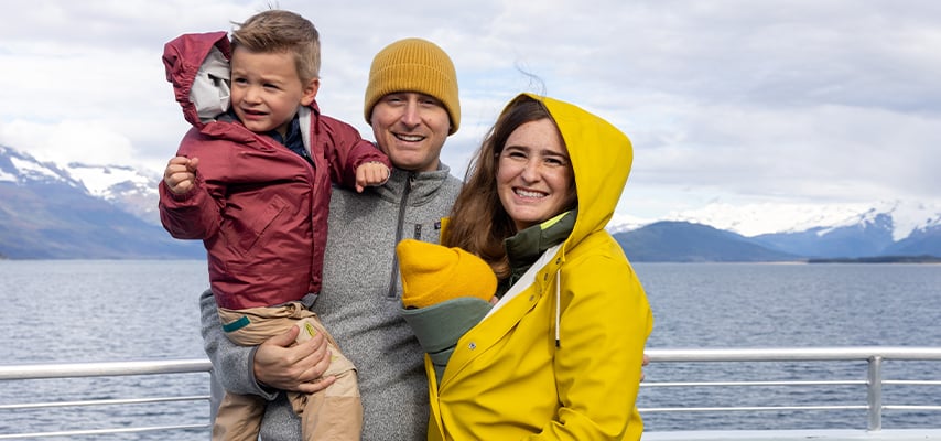 Happy family of three in winter gear smiling on cruise ship deck with Alaskan mountains behind.