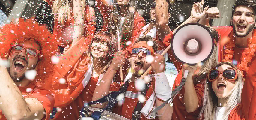 Excited sports fans in red celebrating with confetti falling, one holding a megaphone, all cheering with arms raised.