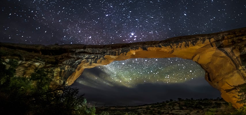 A wide sandstone natural bridge frames a glowing, star-dense night sky at Natural Bridges National Monument, Utah.