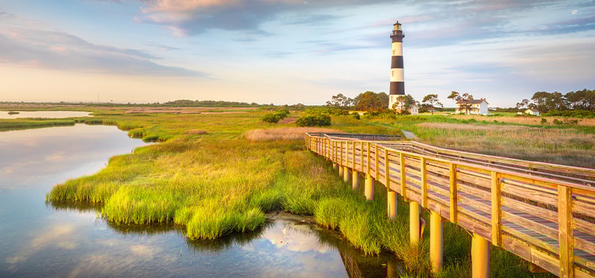Wooden boardwalk through green marsh leading to black and white striped lighthouse