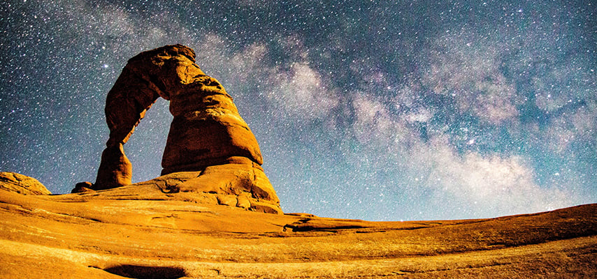Delicate Arch silhouetted against a vivid blue night sky with the Milky Way streaming behind it at Arches National Park, Utah.