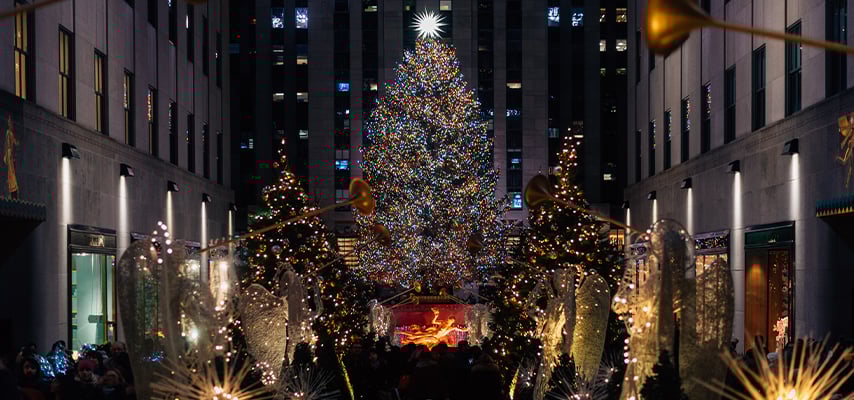 Massive Christmas tree and decorations illuminate Rockefeller Center plaza surrounded by skyscrapers.