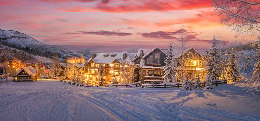 Snow-covered mountain lodge buildings glow warmly against pink sunset sky and snowy landscape.