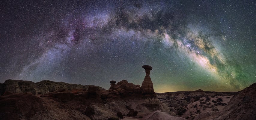 A full arc of the Milky Way in purple and green stretches over a toadstool hoodoo rock formation in Utah's dark desert landscape.